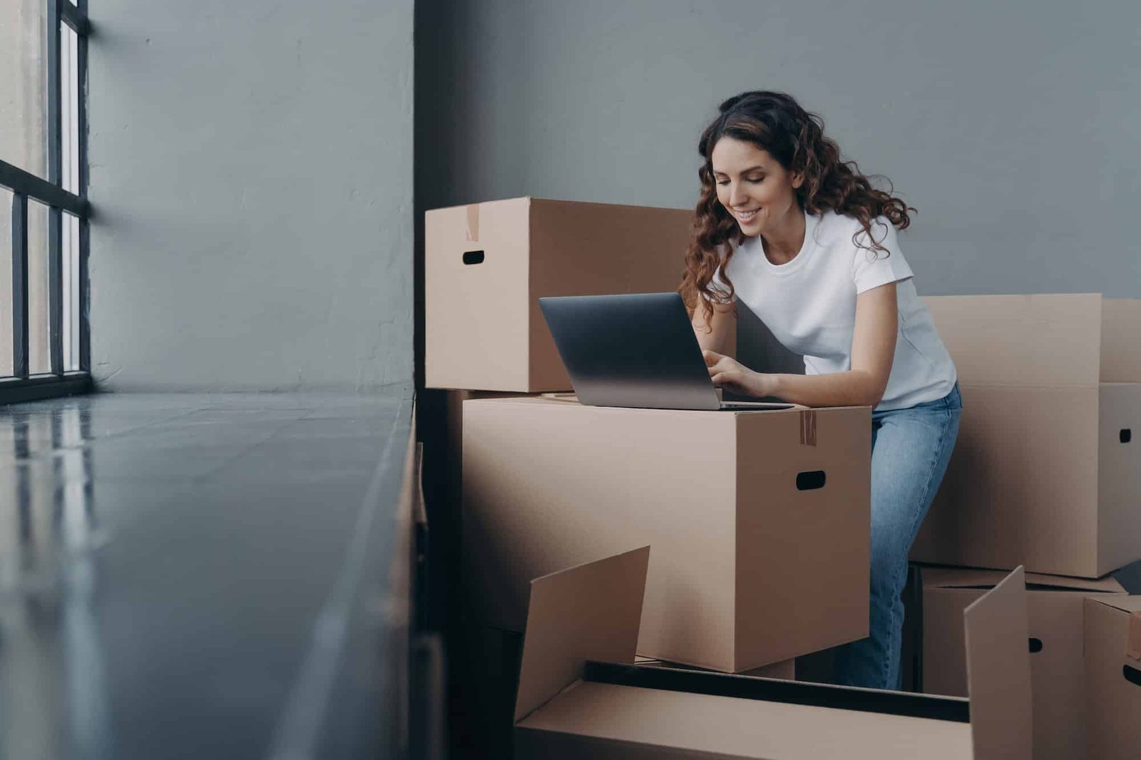 Female with boxes works at laptop, orders moving company, shopping online. Ecommerce, relocation day