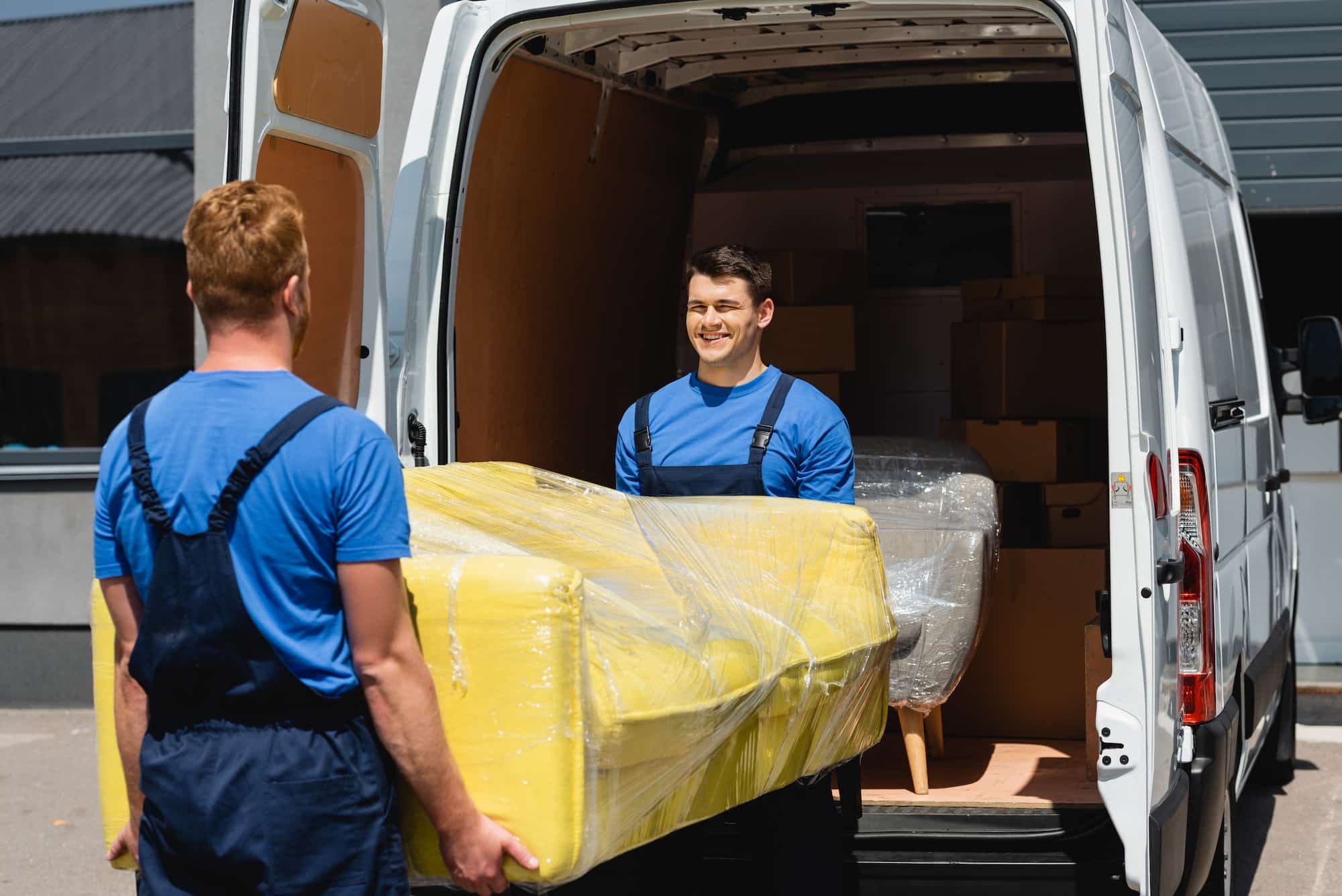 Selective focus of movers holding sofa in stretch wrap near truck on urban street